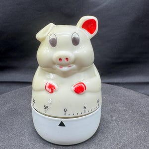 May include: A vintage kitchen timer shaped like a pig. The pig is off-white with red accents on the ears, snout, and hands. The timer has a white base with a black triangle and numbers indicating minutes.