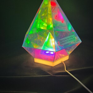 May include: A multi-colored iridescent pyramid lamp with a white base and a white cord. The lamp is lit and casting a colorful glow. The lamp has a sticker on it that says "Tips: Please tear off the protective film before use."
