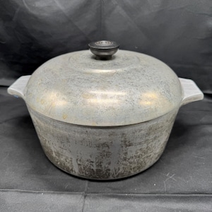 May include: A vintage aluminium cooking pot with a lid and a black knob. The pot has two side handles and a worn, textured surface. The pot is round and appears well-used.