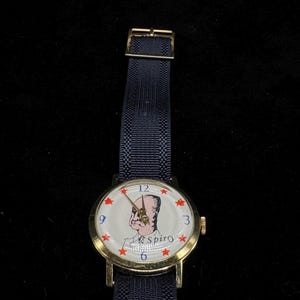 May include: A gold-toned wristwatch with a navy blue fabric band. The watch face features a portrait and the name "Spiro" with red star hour markers. The watch band is approximately 0.75 inches wide.