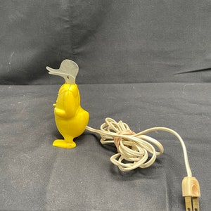 May include: A yellow plastic duck nightlight with a gray beak and a white cord. The duck is plugged into an electrical outlet.