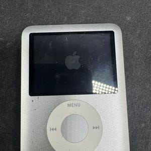 May include: Silver iPod with a black screen showing the Apple logo. The device features a circular control pad with a menu button and navigation controls. The iPod is connected to a white cable.