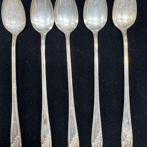 Pode incluir: Cinco colheres de prata dispostas em fila. Cada colher tem uma tigela oval e um cabo longo e fino. As alças apresentam um desenho floral perto da base. As colheres são exibidas em um fundo escuro.