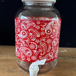 May include: A clear glass beverage dispenser featuring a wide red band with a white paisley and floral pattern wrapped around the center. The container includes a white plastic lid and a white plastic spigot attached to the lower front section.