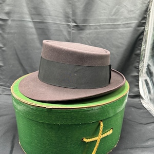Op de afbeelding: Een donkergrijze fedora-hoed met een zwarte band, die in een groene hoedenbox met een bruin touwhandvat ligt.