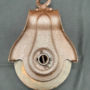 May include: A rusty cast iron pulley with a wooden wheel. The pulley has a loop at the top for hanging and a metal pin in the center.