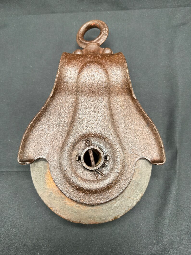May include: A rusty cast iron pulley with a wooden wheel. The pulley has a loop at the top for hanging and a metal pin in the center.