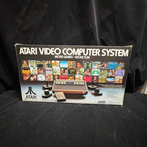 May include: A rectangular cardboard box for an Atari Video Computer System, model number CX-2600A. The box features the Atari logo, the text "MORE GAMES-MORE FUN," and a collage of various video game screenshots, along with the console and controllers.