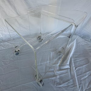 May include: Clear acrylic side table with a unique, angular design. The table has a two-tiered structure and is mounted on small, clear casters for easy mobility. The transparent material allows for a modern and minimalist aesthetic, suitable for various interior styles.