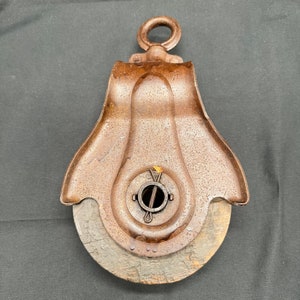 May include: A vintage, rusty metal pulley with a wooden wheel. The pulley has a metal loop at the top and a metal pin in the center of the wheel.