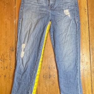 May include: A pair of light blue denim jeans with distressed details. The jeans have a straight leg fit and are measured with a yellow tape measure.