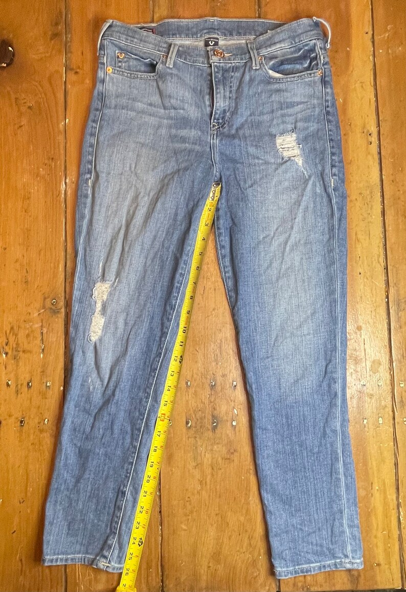 May include: A pair of light blue denim jeans with distressed details. The jeans have a straight leg fit and are measured with a yellow tape measure.