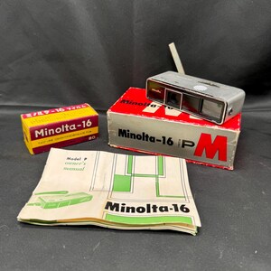 Può includere: Una macchina fotografica vintage Minolta-16 P con corpo argentato e obiettivo nero. La macchina fotografica è appoggiata su una scatola rossa con la scritta "Minolta-16 P" stampata su di essa. C'è anche una scatola di pellicola Minolta-16 e un manuale per la macchina fotografica.