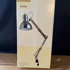 May include: A silver desk lamp with a clamp base. The lamp has a satin steel finish and is functional and stylish. The box reads "Innovations by Meijer" and "Metro lamp".