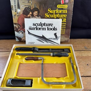 May include: A yellow and white box with a wood carving kit. The box has a picture of a family on the front and the text "Surform Sculpture Kit" and "sculpture surform tools". The kit includes a wood block, a saw, a rasp, and a file.