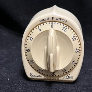 May include: A cream-colored kitchen timer with a central rotating knob. The face features black markings for 0 to 55 minutes, with the text "MINUTE MINDER" at the top and "Robertshaw Lux" printed in black script at the bottom base.