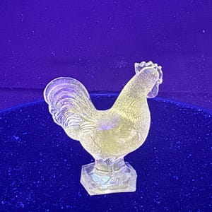 May include: A clear, glass rooster figurine with a textured surface. The rooster is standing on a hexagonal base and has a detailed comb and tail feathers. The figurine is set against a dark blue background.