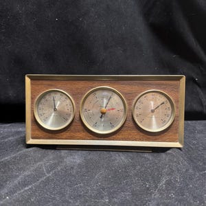 Könnte beinhalten: Eine rechteckige Wetterstation mit einem Rahmen in Holzoptik und einem messingfarbenen Rand. Sie enthält drei kreisförmige analoge Anzeigen: ein Thermometer links, ein Barometer in der Mitte und ein Hygrometer auf der rechten Seite.