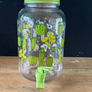 May include: A clear glass beverage dispenser featuring a repeating pattern of various green cacti illustrations. The container includes a matching lime green screw-on lid and a lime green plastic spigot located at the bottom front for dispensing.