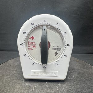 May include: A white kitchen timer with a black central knob and a circular dial marked from 0 to 55 minutes. The face features red text reading TO START, TURN PAST 55 MIN. THEN... and black text reading BACK TO DESIRED TIME. Everyday Living is printed below.