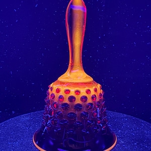 Puede incluir: Una campana de cristal con patrón de puntos en relieve y tono rojo profundo que brilla con una luminiscencia naranja y amarilla bajo luz ultravioleta. La campana tiene un mango liso y alargado, y una base acampanada con un borde brillante.