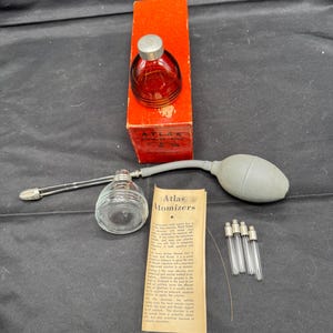 May include: An antique Atlas Atomizers set, including a red glass bottle with a silver cap, a clear glass atomizer, a rubber bulb, and a paper instruction sheet. The set also includes several glass tubes. The items are on a dark surface.