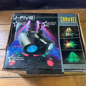 Op de afbeelding: Een zwarte J-FIVE dubbele roterende LED-lichteffecteenheid met twee blauwe lenzen. De doos is voorzien van het CHAUVET-logo en de tekst en geeft aan dat de output vergelijkbaar is met twee 100W halogeenlampen. Het ontwerp is compact met volledige kamerdekking.