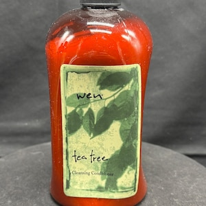 May include: A red bottle of Wen tea tree cleansing conditioner with a black cap. The label features green leaves and the words "wen" and "tea tree". The bottle has a curved shape.