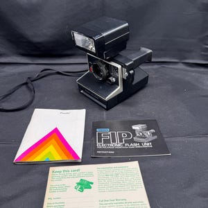 Op de afbeelding: Een vintage Polaroid Pronto! camera met een zwarte behuizing, zilveren afwerking en een flitser. De afbeelding toont ook instructiehandleidingen en een garantiekaart. De camera heeft een lens en een handvat. De flitser is zwart.