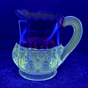 Puede incluir: Una pequeña jarra de cristal transparente con una base decorativa y un borde rojo oscuro. La jarra tiene un asa y un pico. Las palabras "Souvenir of CYPRUS" son visibles en la parte delantera.