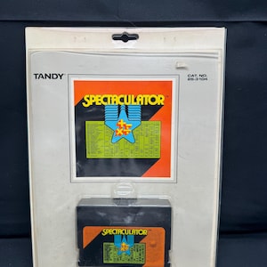 Könnte beinhalten: Eine Tandy Spectaculator Videospielkassette in ihrer Originalverpackung. Die Kassette ist schwarz mit einem orangefarbenen Etikett, auf dem "Spectaculator" steht. Die Verpackung ist weiß mit einem rot-schwarzen Etikett, auf dem "Spectaculator" steht, und enthält eine Liste von Spielen.