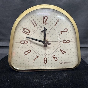 May include: Vintage Gilbert clock with a gold-tone metal frame and a cream-colored face. The clock face features brown numerals and a dotted pattern. The clock hands are black. The clock is marked "Gilbert" and "Made in U.S.A."