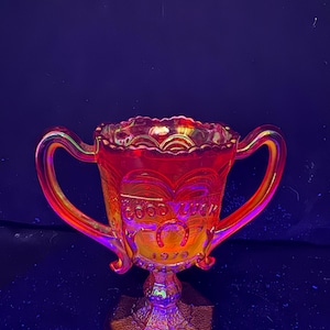 Puede incluir: Una copa trofeo de vidrio de carnaval naranja iridiscente con dos asas, un borde festoneado y una base hexagonal. El frente presenta el texto en relieve GOOD LUCK sobre un símbolo de herradura y el año 1979, sobre una superficie de vidrio texturizada.
