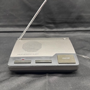 Peut inclure: Une radio grise Weatheradio Alert avec une antenne télescopique. La radio comporte une zone de haut-parleur perforée et des boutons étiquetés "ALERT", "TEST" et "WEATHER". Le texte "WEATHERADIO ALERT" est imprimé en haut.