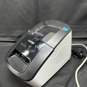 Op de afbeelding: Een Brother QL-700 labelprinter in zwart en wit. Het heeft een doorzichtige bovenkant, bedieningsknoppen en een labeluitvoersleuf. Een netsnoer is aangesloten. De printer is ontworpen voor het maken van labels.