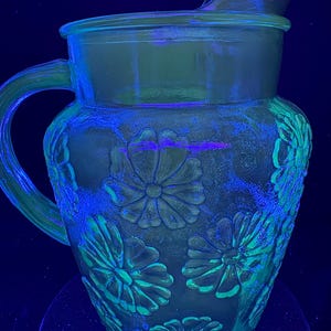 May include: A green glass pitcher with a handle and a lid. The pitcher is decorated with embossed floral patterns. The image is illuminated with a blue and green light, highlighting the details of the glass and the floral design.