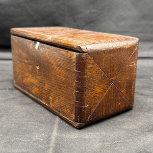 Könnte beinhalten: Eine rechteckige Holzkiste mit Scharnierdeckel. Die Kiste besteht aus dunkelbraunem Holz mit sichtbaren Maserungsmustern. Die Ecken sind mit einem kreuzförmigen Design verbunden. Die Kiste scheint Vintage oder antik zu sein.