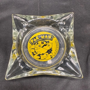 May include: A clear glass ashtray with a square shape and a circular center. The center features a yellow and black Pac-Man design with the text "PAC-MAN". The ashtray has a vintage aesthetic.