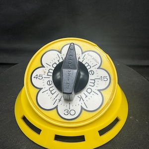 May include: A yellow kitchen timer with a black dial and a white face featuring the word "time mark". The timer has a flower-shaped design with numbers indicating minutes. The base has cut-out slots.