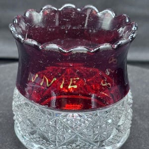 Op de afbeelding: Een kleine, heldere glazen beker met een geslepen glazen voet en een dieprode, geschulpte rand. Het rode gedeelte heeft de naam "Annie" in gouden letters geëtst. De beker is waarschijnlijk vintage en decoratief.