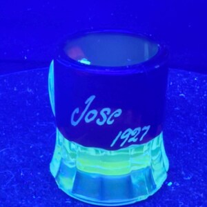Op de afbeelding: Een klein, helder glas met een donkerblauwe band en een lichtgroene basis. De naam "Jose" en het jaar "1927" staan in witte letters op de blauwe band. Het glas heeft een unieke, uitlopende basis.