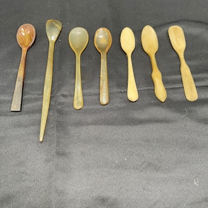 May include: A set of eight spoons made from horn. The spoons are in various shapes and sizes, with some having a rounded bowl and others having a more pointed shape. The spoons are all a light brown color.