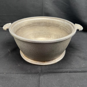 Puede incluir: Una olla de aluminio plateada con dos asas y una superficie texturizada. La olla tiene una tapa.