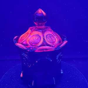 Puede incluir: Un tarro de vidrio oscuro con tapa y diseño decorativo. El tarro es negro con un brillo rosa y naranja, y la tapa tiene un remate puntiagudo. El tarro está sobre una superficie oscura sobre un fondo azul.