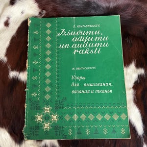 May include: A green book titled "Izšuvumu, adijumu un audumu raksti" with text in a foreign language. The book cover features decorative patterns and text in white. The book is lying on a brown and white patterned surface.