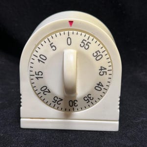 May include: An off-white kitchen timer with a circular dial featuring black numbers from 0 to 55 in five-minute increments. A central vertical knob is used for setting the time, and a small red triangle indicator is positioned at the top.