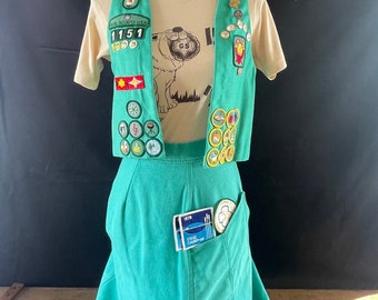May include: A green Girl Scout uniform with a vest and skirt. The vest and skirt are decorated with badges and patches. The uniform also includes a green beret with a Girl Scout logo.