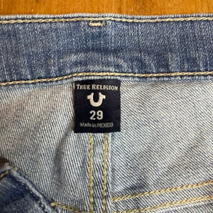 May include: A blue denim jean label with the text "True Religion" and a stylized "U" logo. The label also includes the size "29" and the text "Made in Mexico."