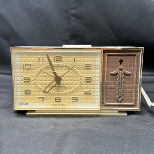 Könnte beinhalten: Ein goldfarbener analoger Wecker mit einer braunen Kunststoff-Seitenteil. Das Zifferblatt hat einen weißen Hintergrund mit schwarzen Zahlen und Zeigern. Auf dem Zifferblatt steht auch der Text "Westclox Electric" und "dialite". Die Uhr ist in den USA hergestellt.