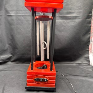 May include: A red and black electric wind chime device featuring a rectangular red base and top connected by four black pillars. Silver metal chime tubes hang from the center, with a central ring and a black power cord extending from the base.